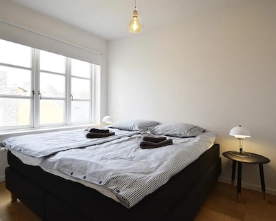 Soveværelse med dobbeltseng, gråstribet sengetøj, to håndklæder og vindue.