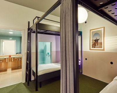 Et lyst soveværelse med køjeseng, grønt tæppe og badeværelse i baggrunden.